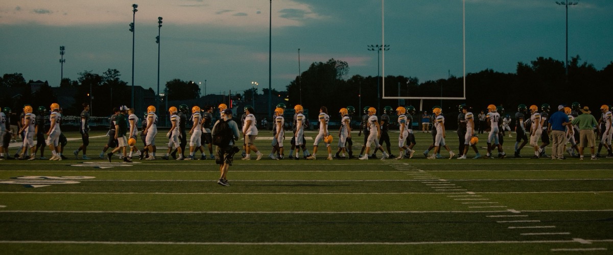 Two youth football teams shaking hands across the field at dusk under the lights